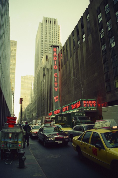 A street in New York City is filled with cars and taxis, with the Radio City Music Hall sign visible on the right.