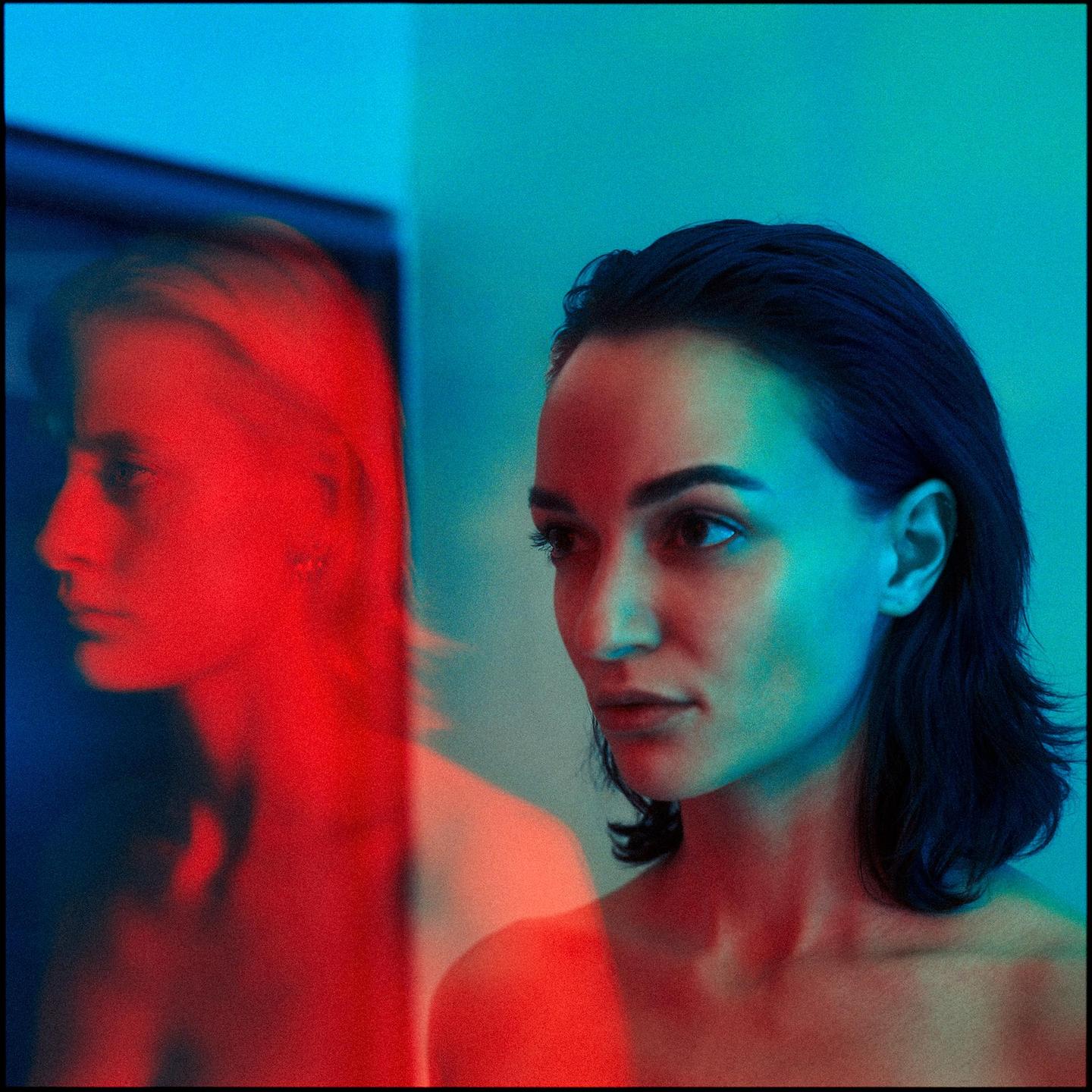 A woman is illuminated by blue and red light, with a red reflection of her face visible on the left.