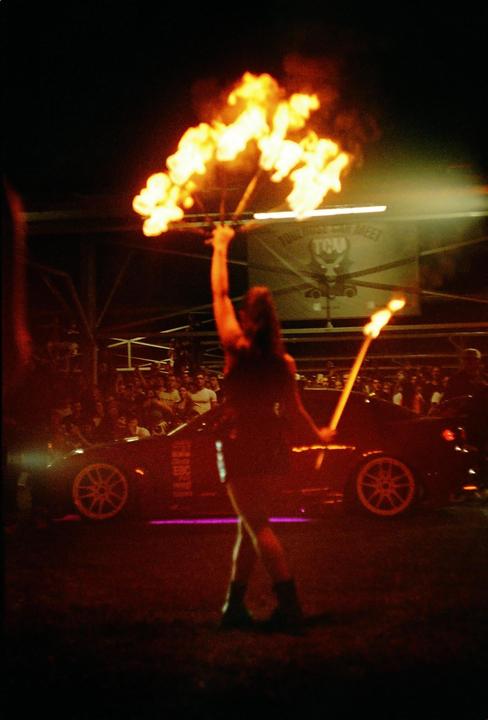 A woman performs with fire in front of a crowd and a car at night.