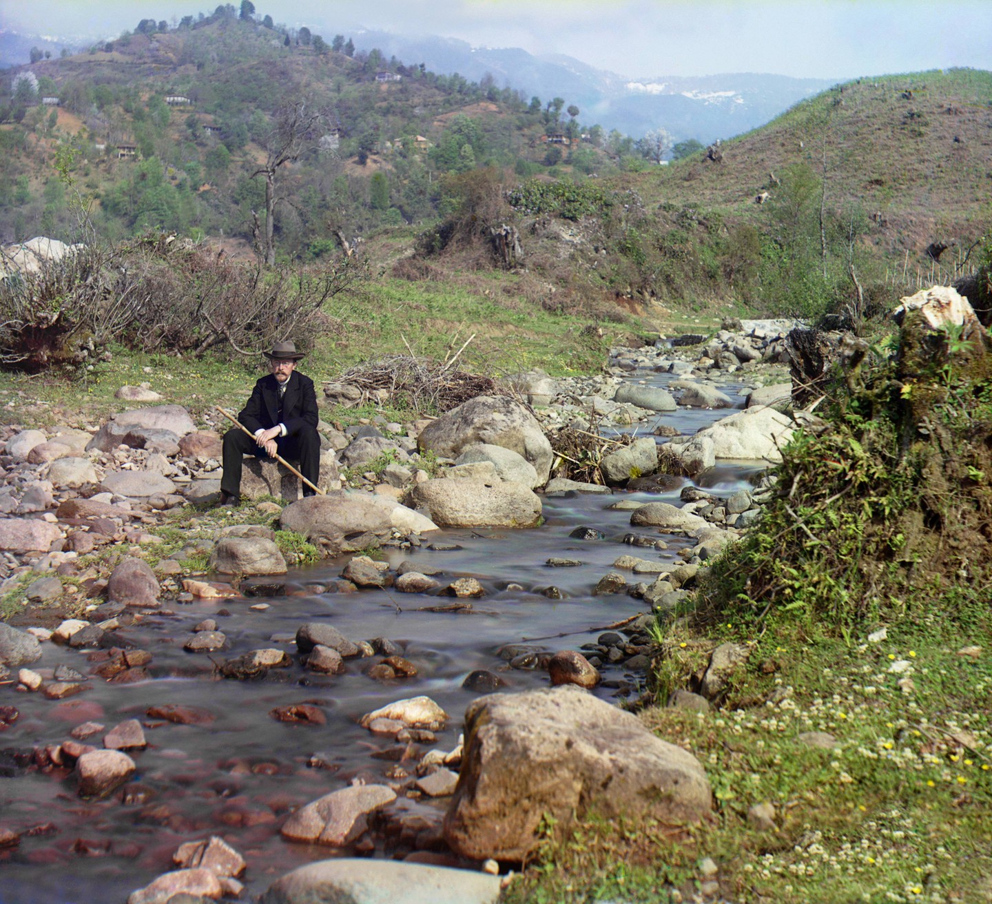 A man in a suit and hat sits by a rocky stream in a mountainous landscape.