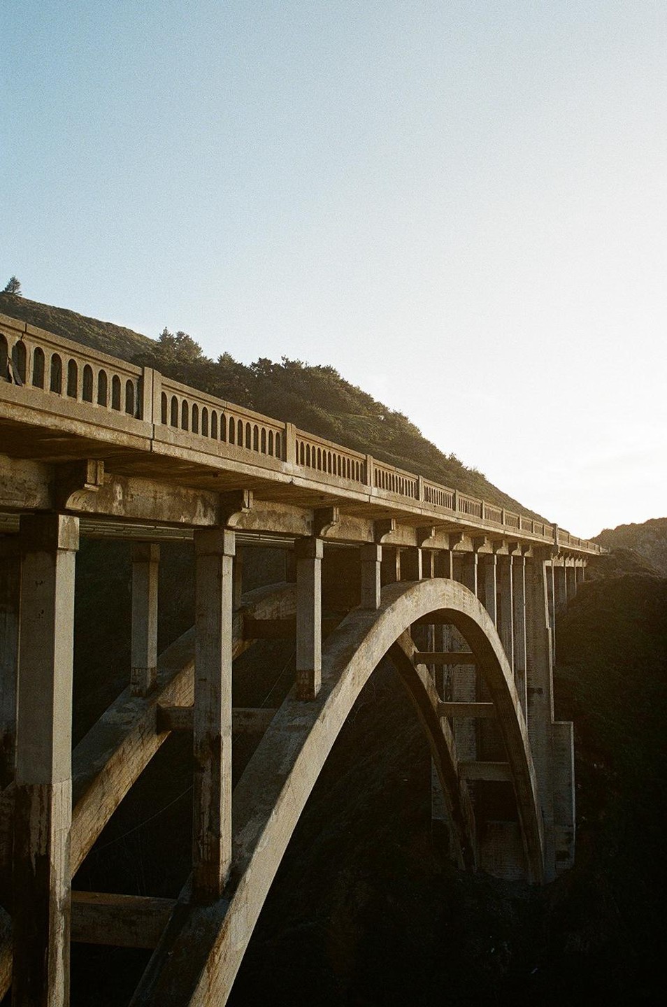 A concrete bridge with arches spans a dark valley, leading to a hillside covered in trees under a bright sky.