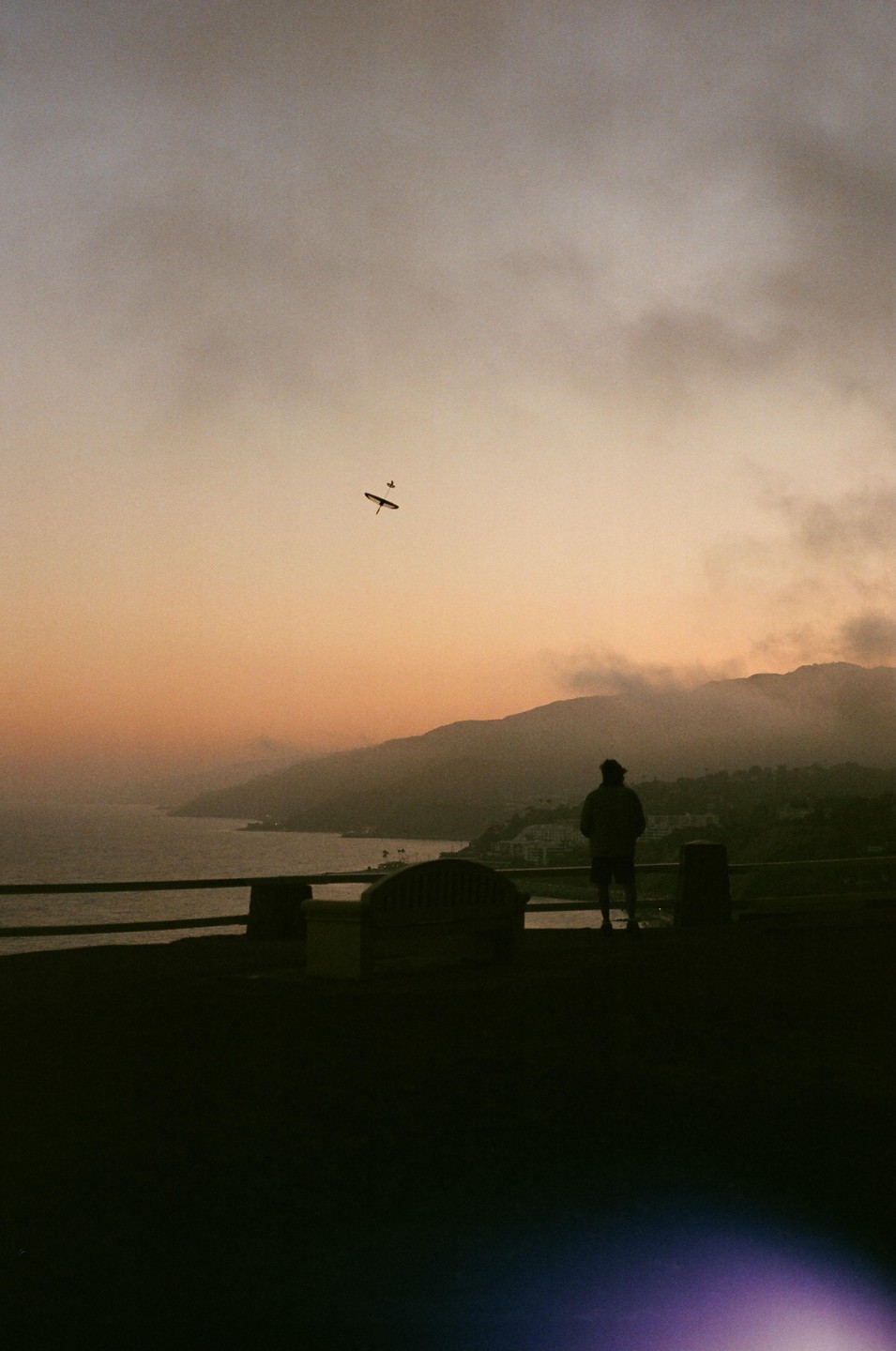 A lone man stands on a hill overlooking a bay with mountains in the distance, as a small plane flies overhead during sunset.