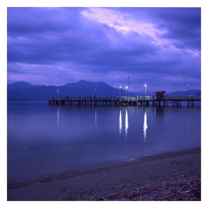 A long pier with lights extends into a calm body of water with mountains in the background under a purple sky.