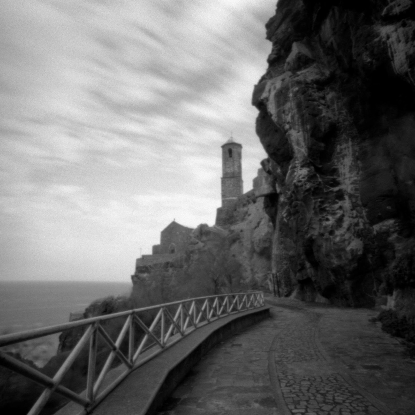 A church with a bell tower stands on a cliff overlooking the ocean, with a paved path and wooden railing in the foreground.