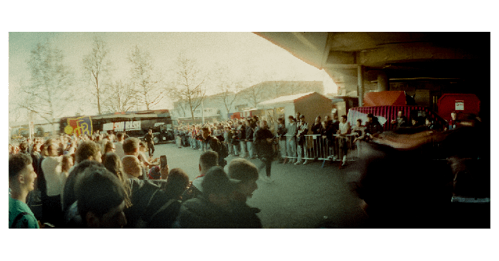 A large crowd of people gathers outdoors, with some standing behind a metal barrier and a bus visible in the background.