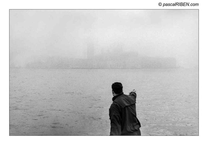 A man points towards a hazy body of water with a faint outline of a city in the distance.