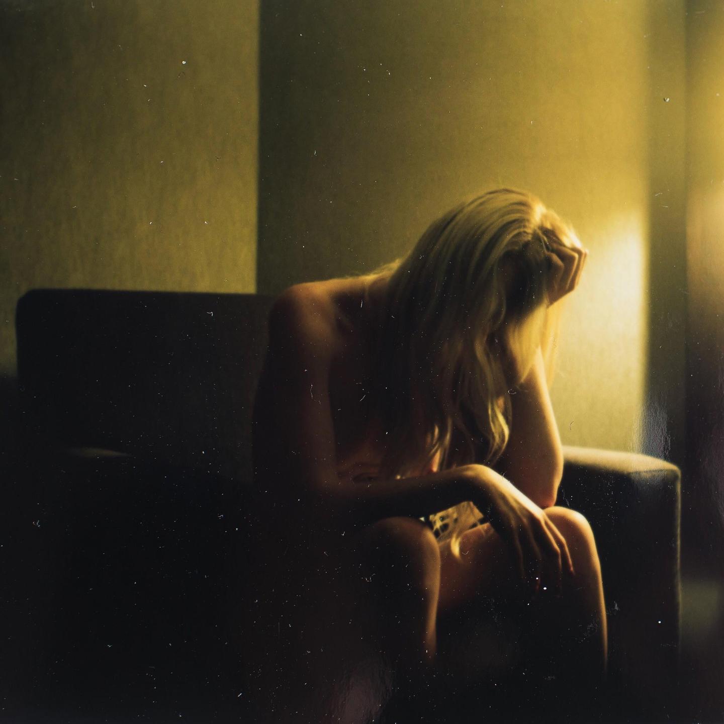 A woman with long blonde hair sits on a chair with her head in her hand.