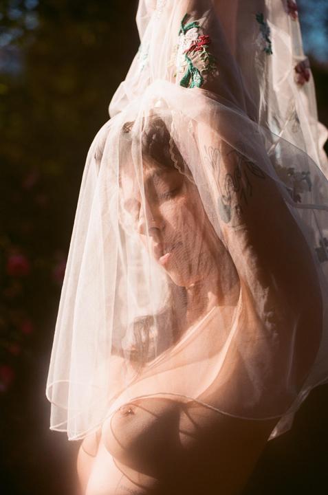 A woman with closed eyes is covered by a sheer veil with embroidered flowers, revealing her bare chest.