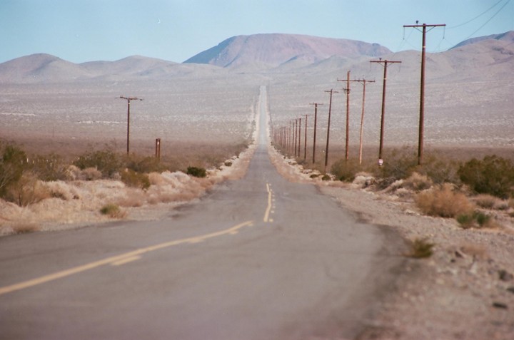 A long, straight road stretches into the distance, flanked by power poles and desert vegetation, with mountains in the background.