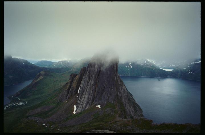 A tall, jagged mountain peak rises from a body of water, surrounded by other mountains under a cloudy sky.
