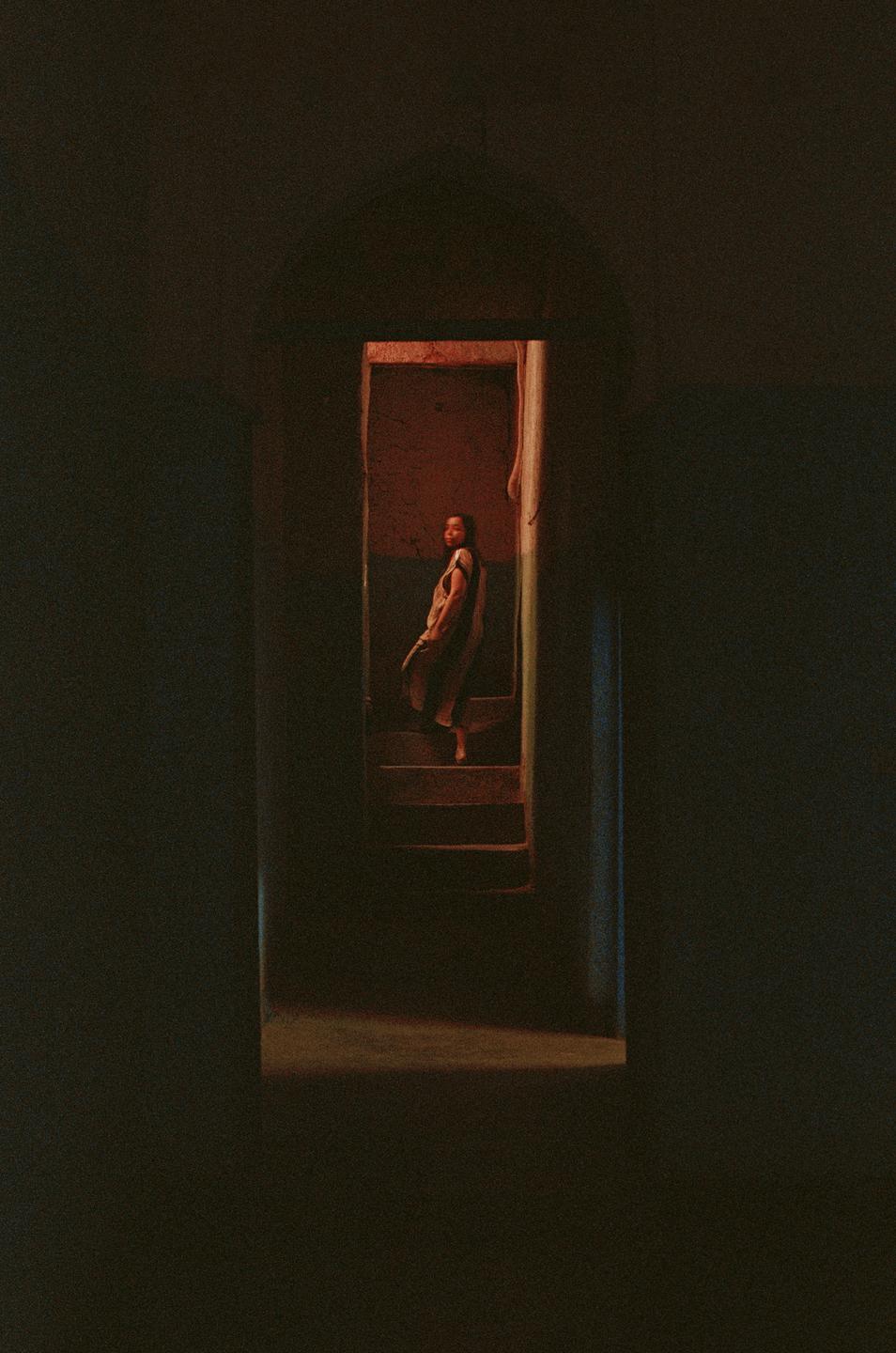 A woman stands on a staircase in a dimly lit room, looking towards the viewer.