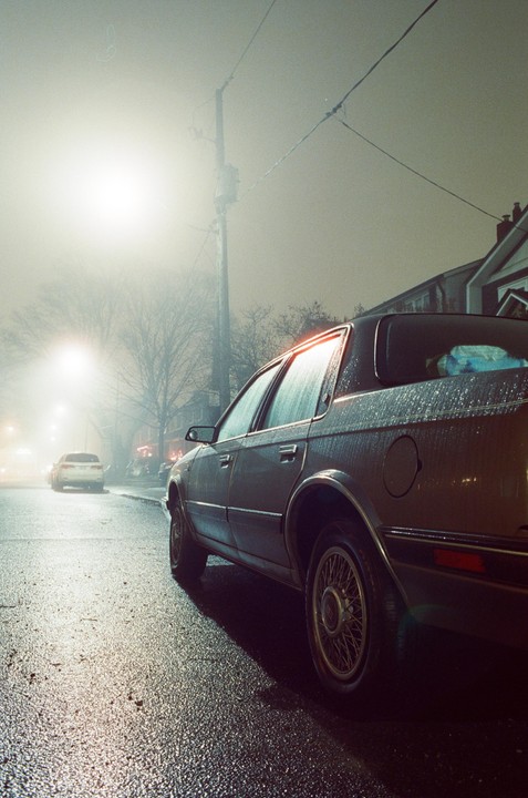 A car is parked on a wet street at night, with another car visible in the distance through the fog.