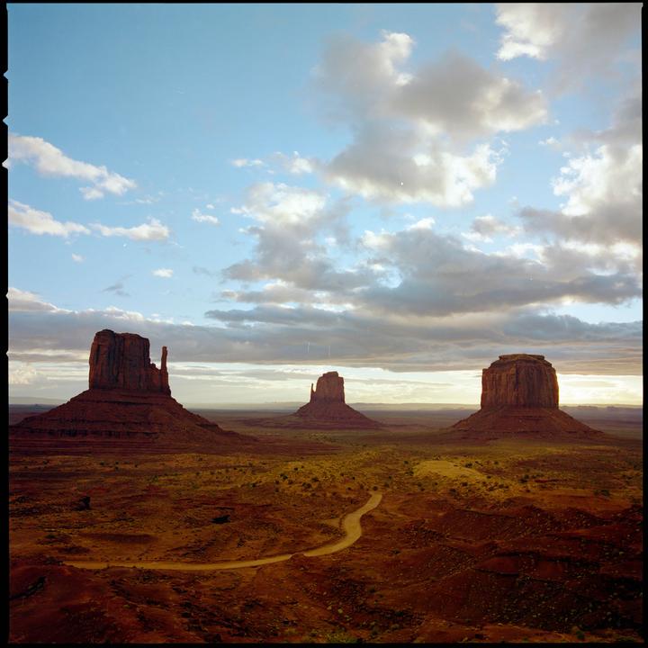 A winding dirt road leads through a desert landscape with three large rock formations under a cloudy sky.