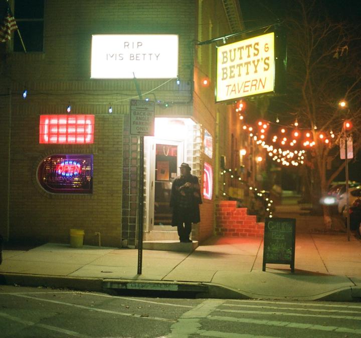 A person stands outside a tavern with glowing signs and string lights at night.