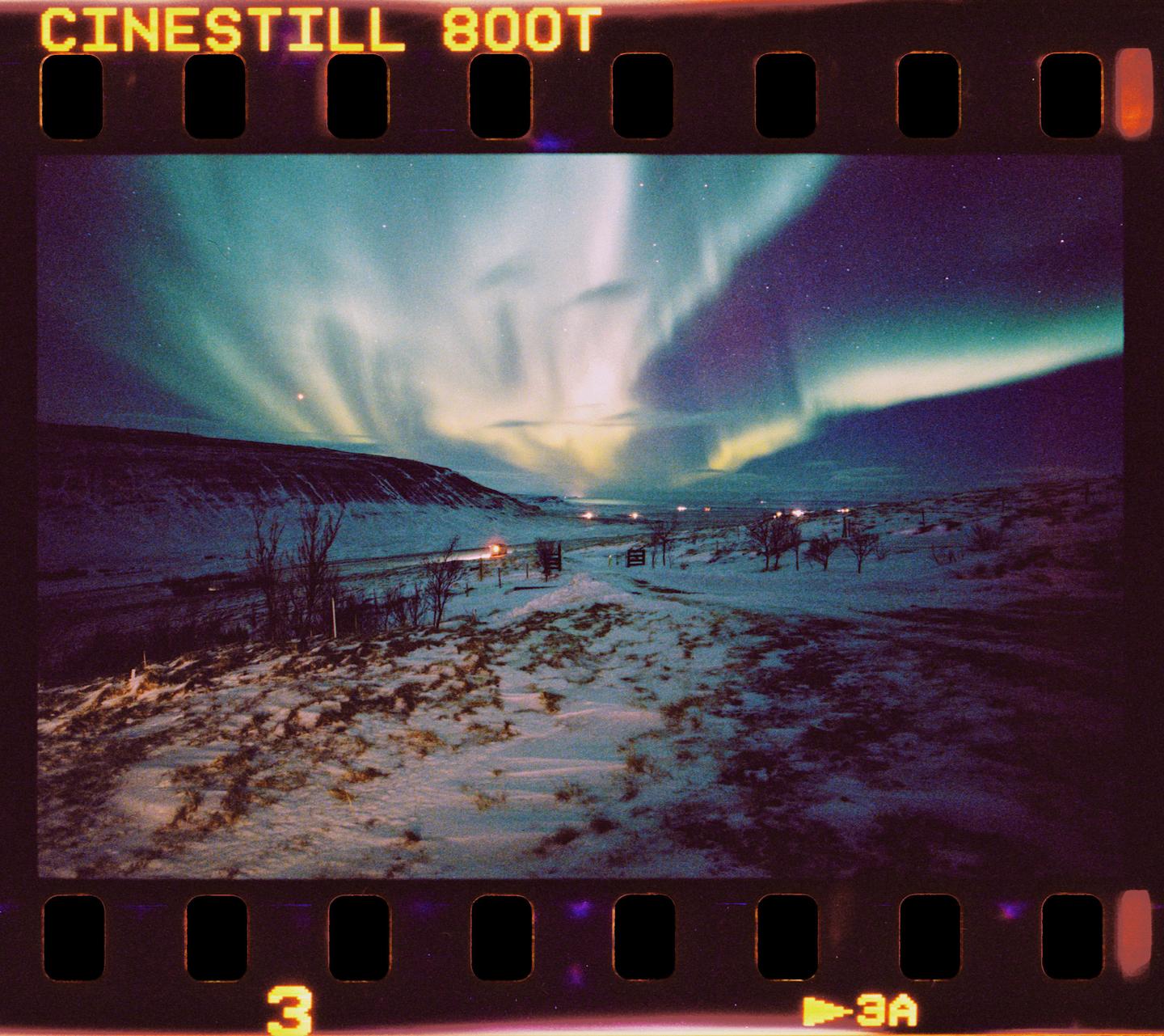 The aurora borealis illuminates a snow-covered landscape with a road and distant lights.