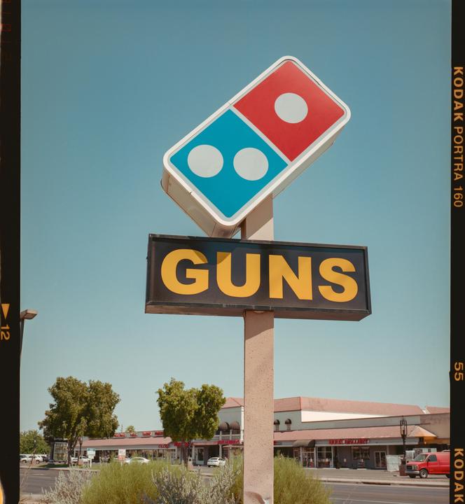 A sign for Domino's Pizza is mounted above a sign that reads "GUNS" next to a street with buildings and trees.