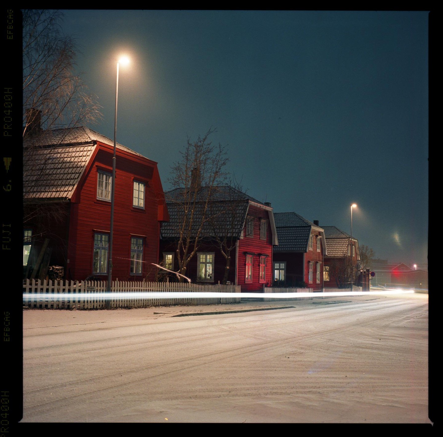 A row of red houses with illuminated windows stands beside a snow-covered street with light trails at night.