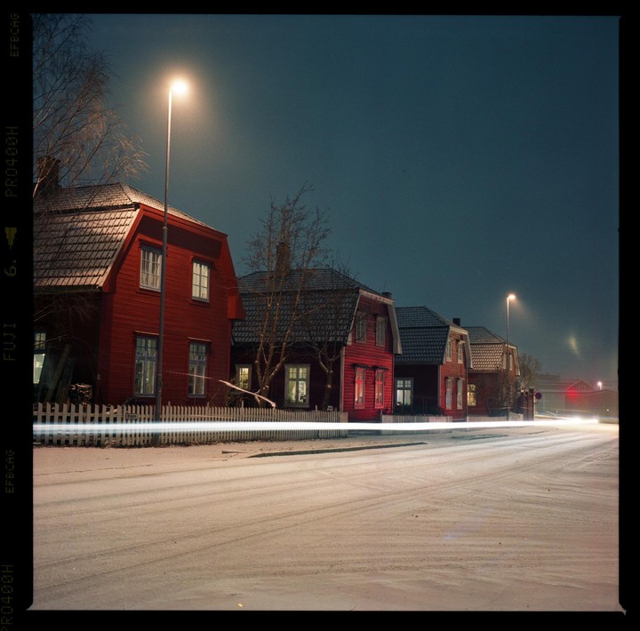 A row of red houses with illuminated windows stands beside a snow-covered street with light trails at night.