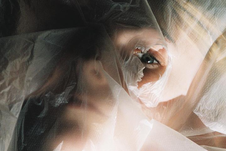 A woman's eye is visible through a torn plastic sheet covering her face.