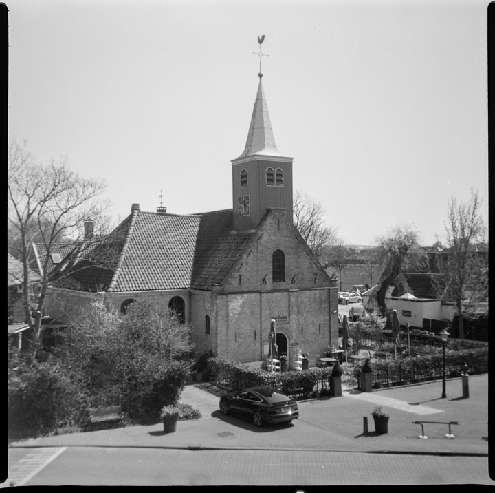 A small church with a tall steeple and a weather vane is surrounded by trees and a street with a car.