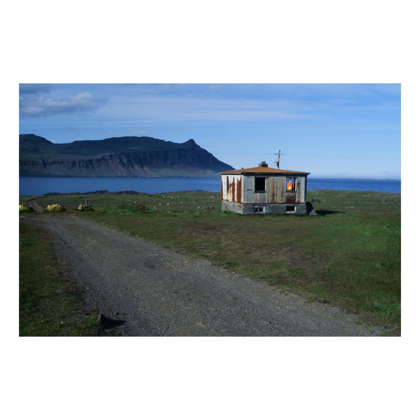 A small, weathered building with a glowing window sits on a grassy shore next to a body of water, with mountains in the distance.