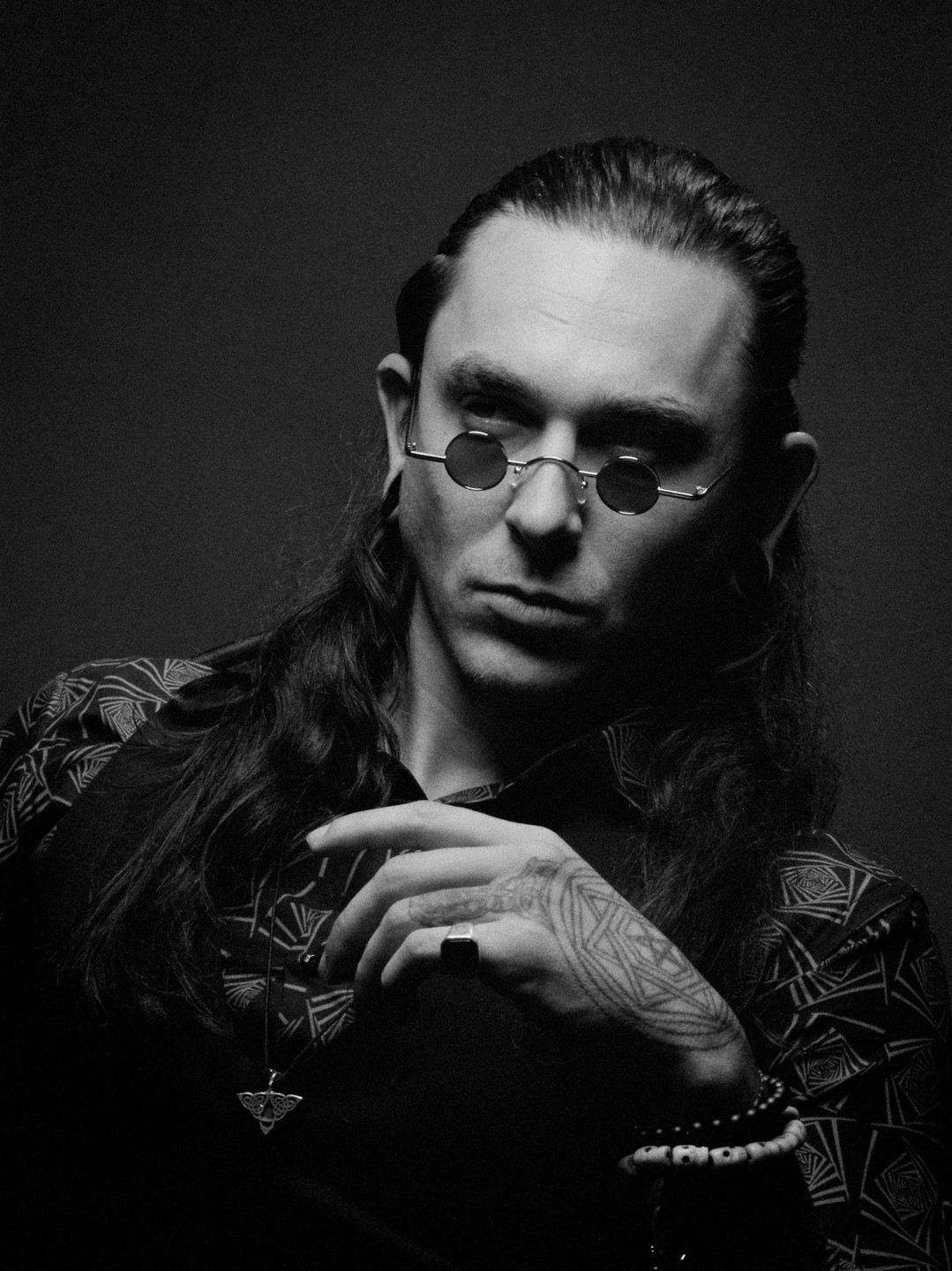 A man with long hair and small round sunglasses looks directly at the viewer.