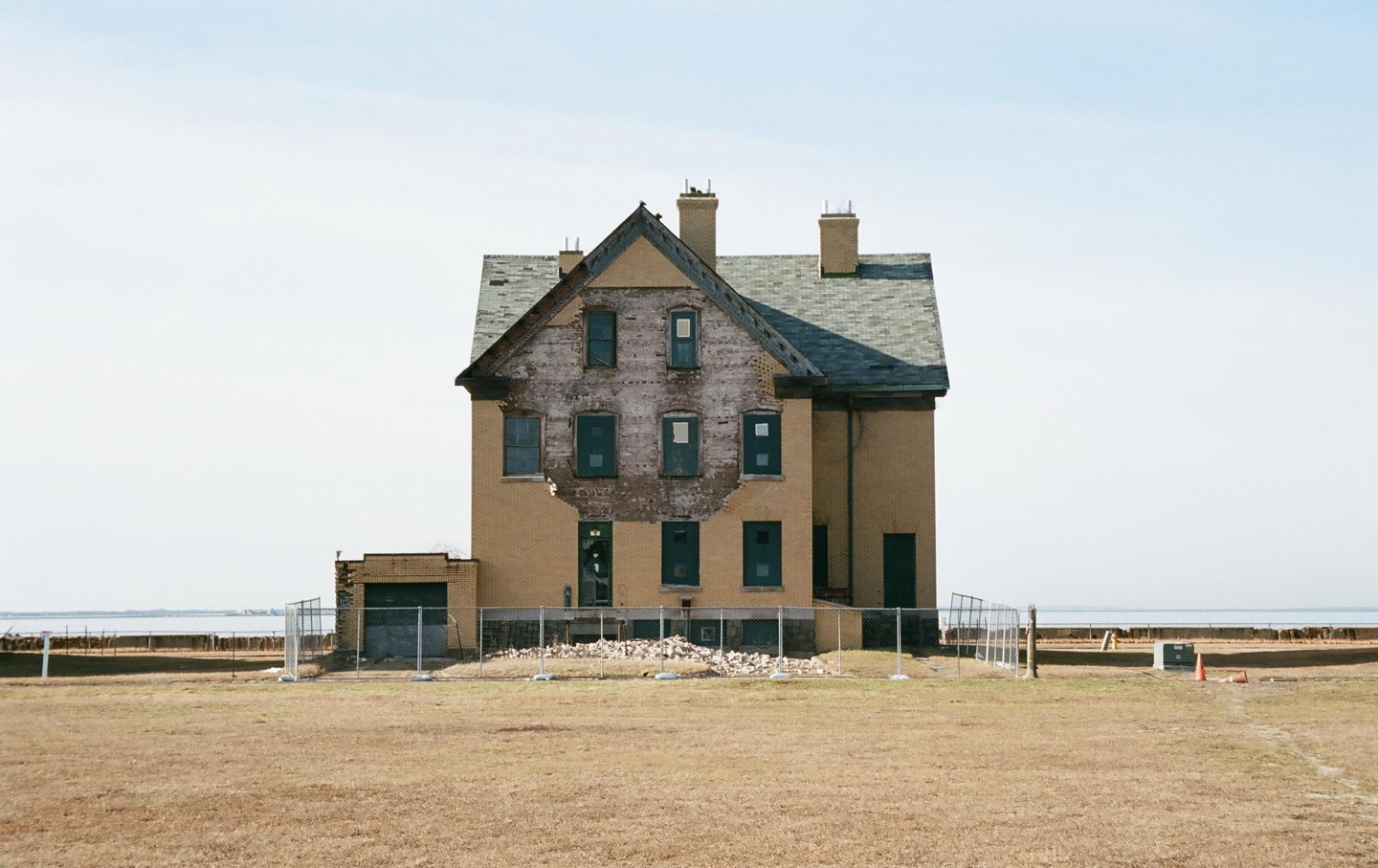A large, abandoned brick house with a damaged facade stands in a dry field next to a body of water.