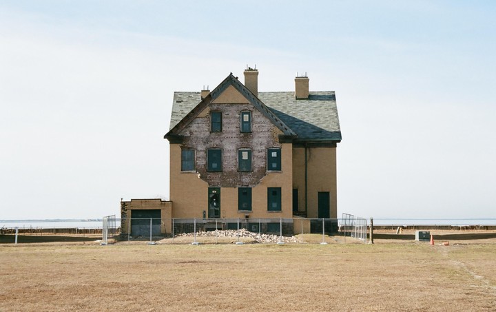 A large, abandoned brick house with a damaged facade stands in a dry field next to a body of water.
