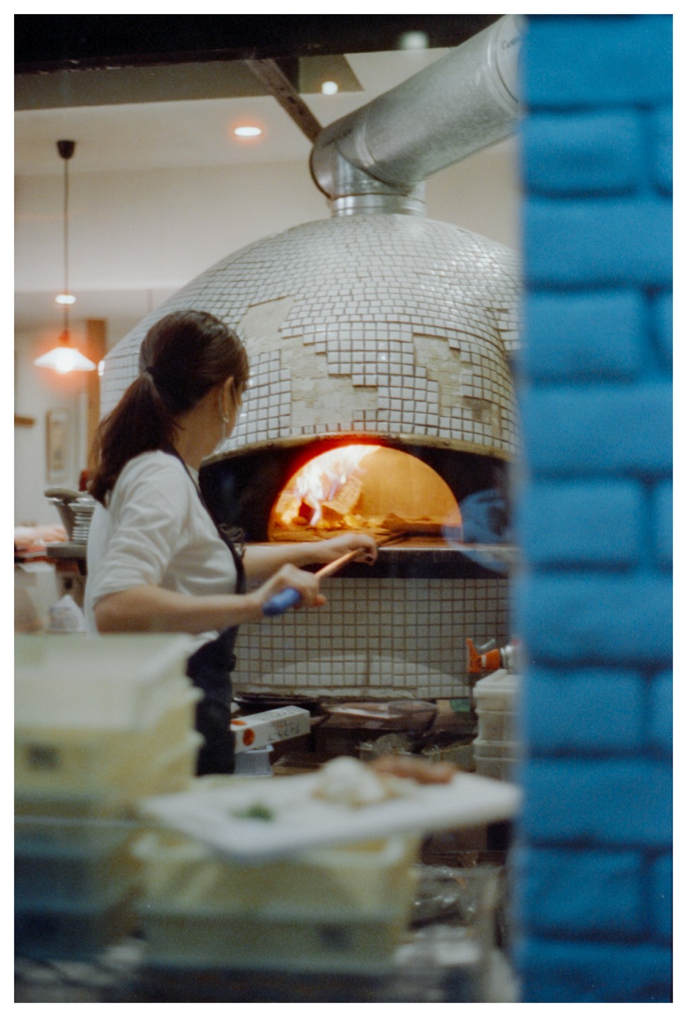 A woman tends to a wood-fired pizza oven in a kitchen.