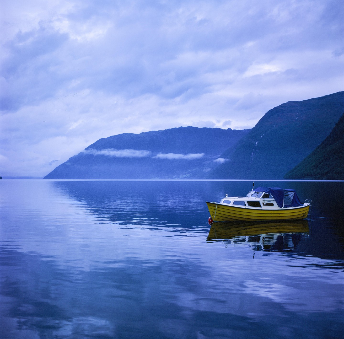 A yellow boat floats on a calm body of water with mountains in the background under a cloudy sky.