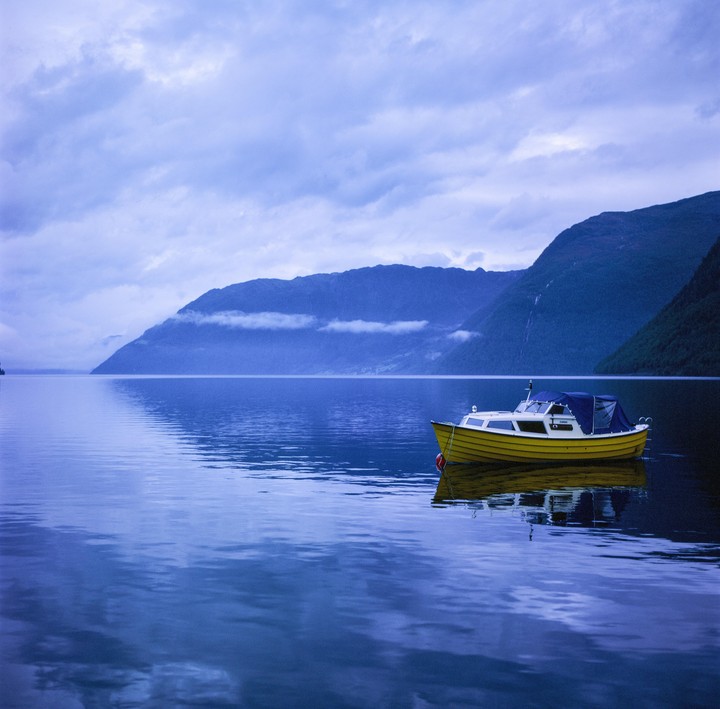 A yellow boat floats on a calm body of water with mountains in the background under a cloudy sky.