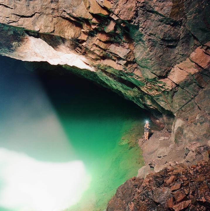 A person with a headlamp stands on a rocky ledge next to a vibrant green body of water inside a cave.