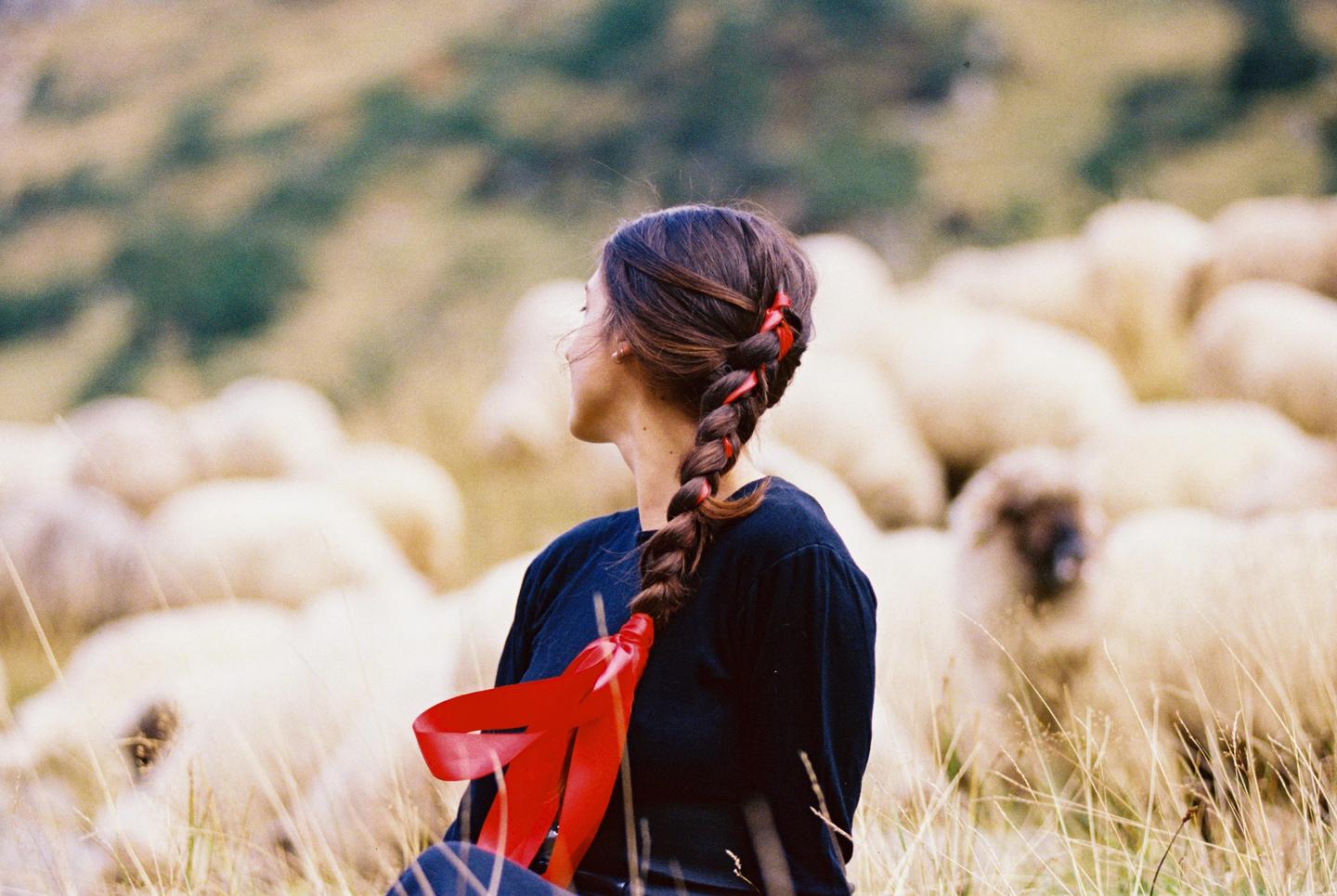 A woman with a long braid adorned with red ribbons sits in a field with sheep in the background.
