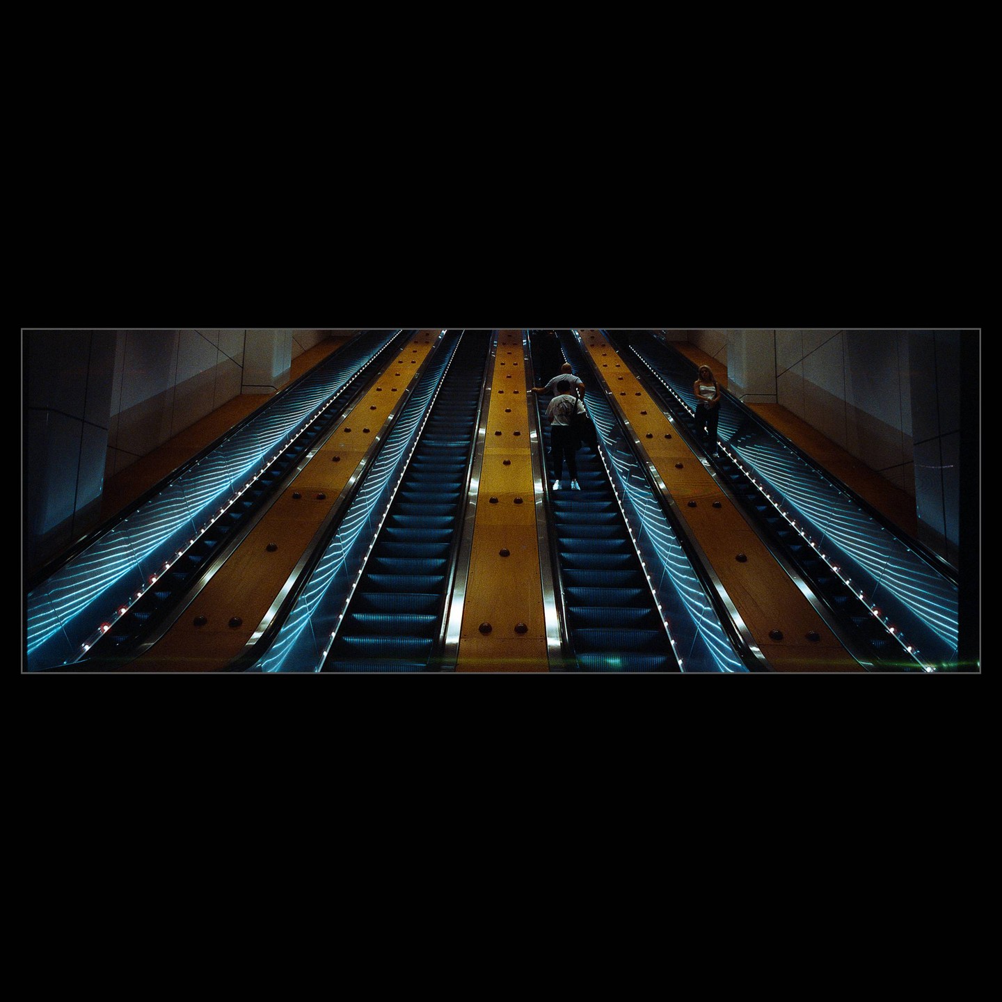 People ride multiple long escalators with blue lights and yellow side panels.