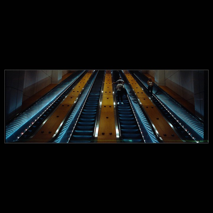People ride multiple long escalators with blue lights and yellow side panels.