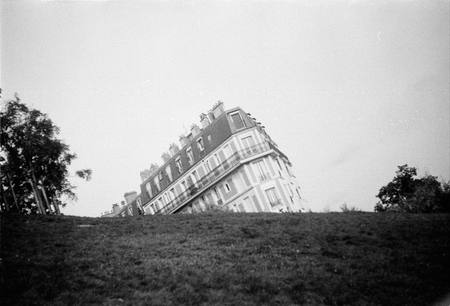 A building with balconies and many windows appears to be sliding down a grassy hill with trees on either side.