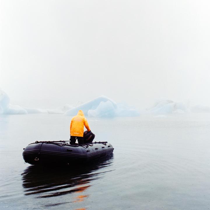 A person in a yellow jacket sits in an inflatable boat on a body of water with icebergs in the distance.