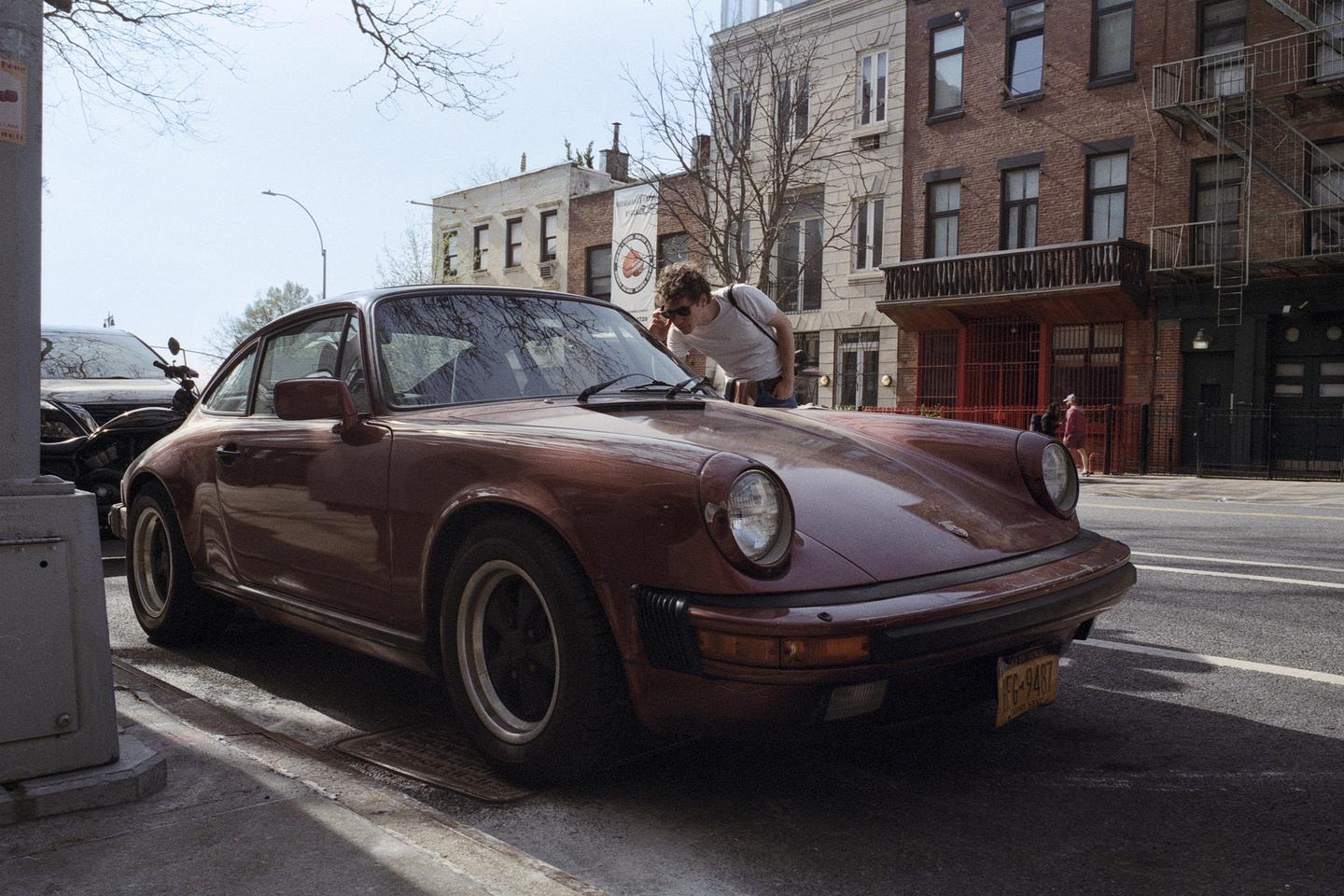 A man wearing sunglasses leans over the hood of a vintage car parked on a city street.