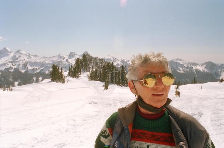 A woman wearing round, reflective sunglasses stands in a snowy mountain landscape with pine trees.
