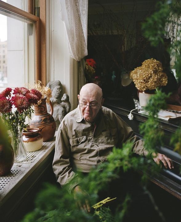 An older man with glasses sits by a window, surrounded by plants and decorative items.