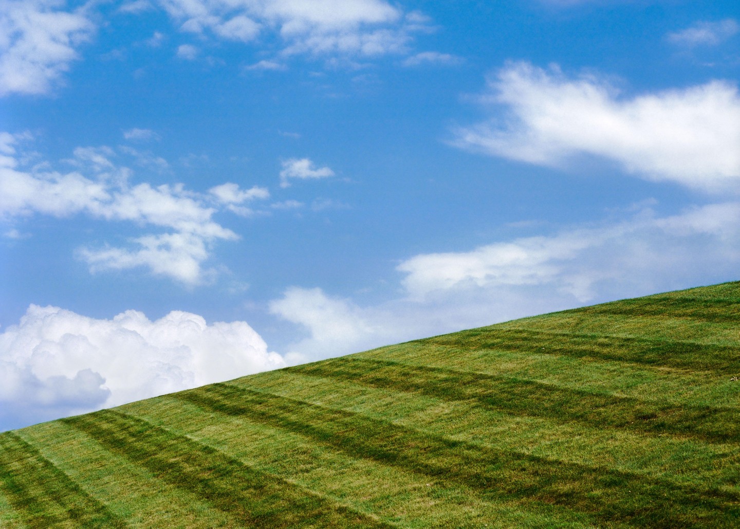 A bright blue sky with white clouds hangs over a green grassy hill with mown stripes.