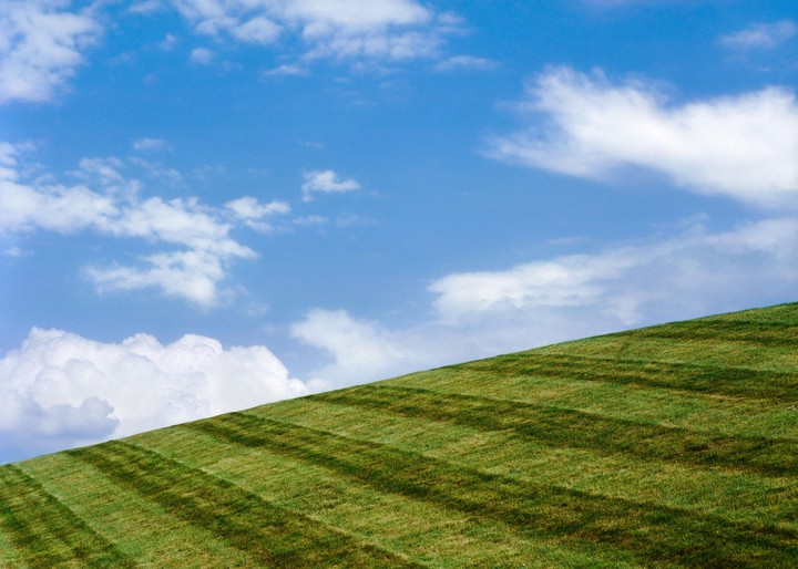 A bright blue sky with white clouds hangs over a green grassy hill with mown stripes.