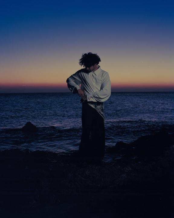 A man stands on a rocky shore with the ocean behind him at dawn.