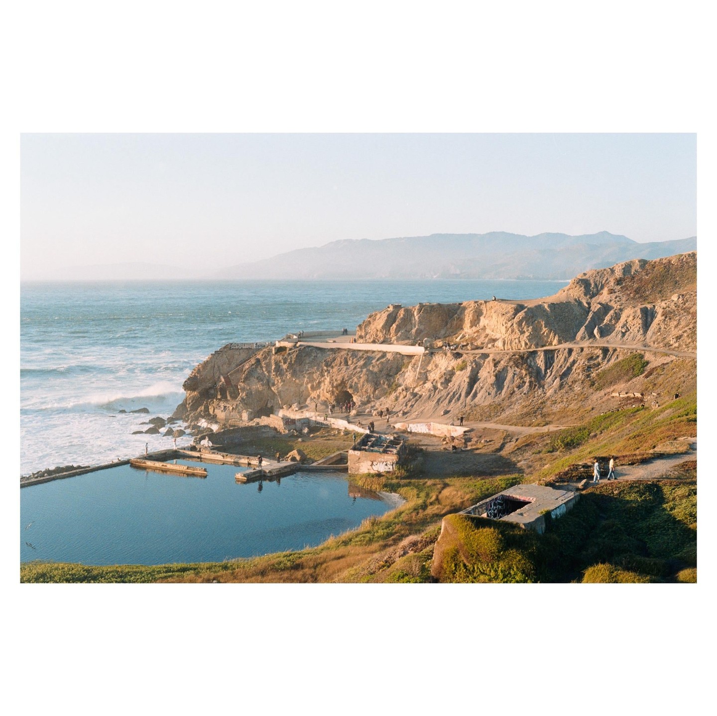 A wide view shows the Sutro Baths ruins and the Pacific Ocean with distant mountains under a clear sky.