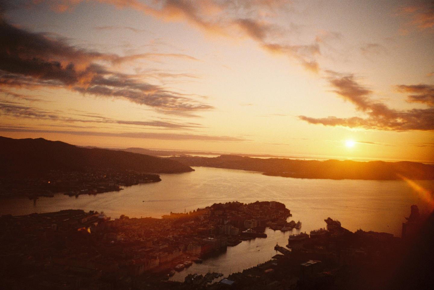 A city and harbor are visible from above at sunset, with mountains in the distance and clouds in the sky.