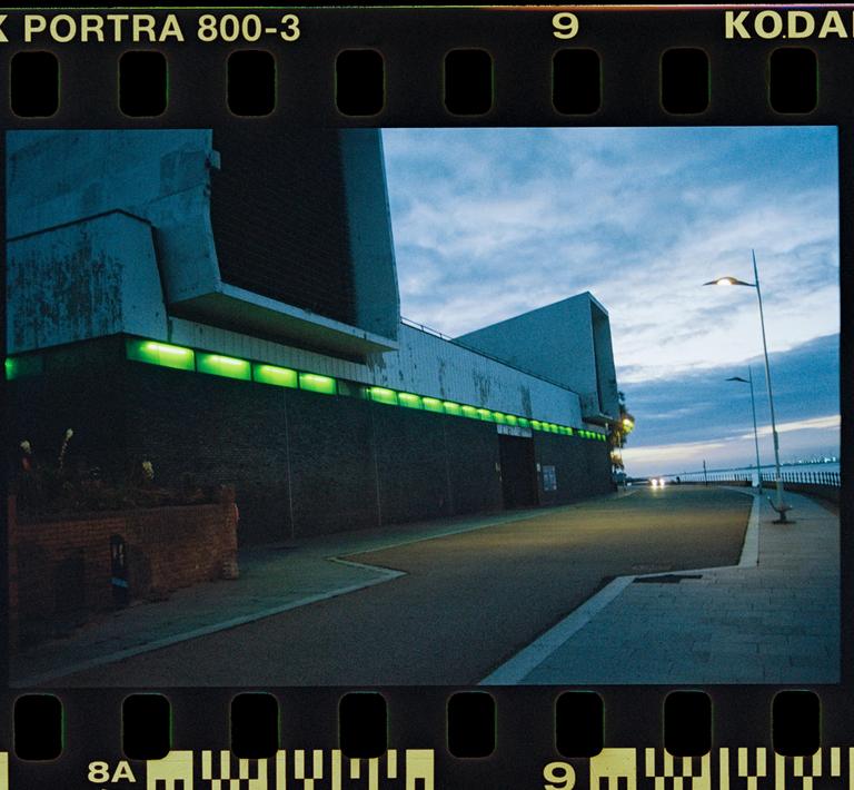 A building with green lights along its facade stands next to a road and a body of water under a cloudy sky.