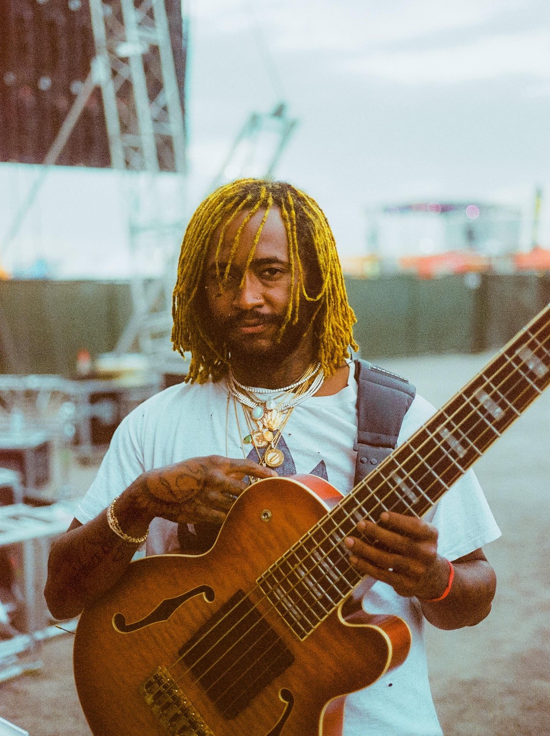 A man with yellow dreadlocks and many necklaces holds a bass guitar.