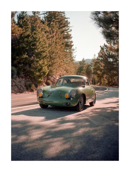 A green vintage car is parked on the side of a winding road surrounded by pine trees.