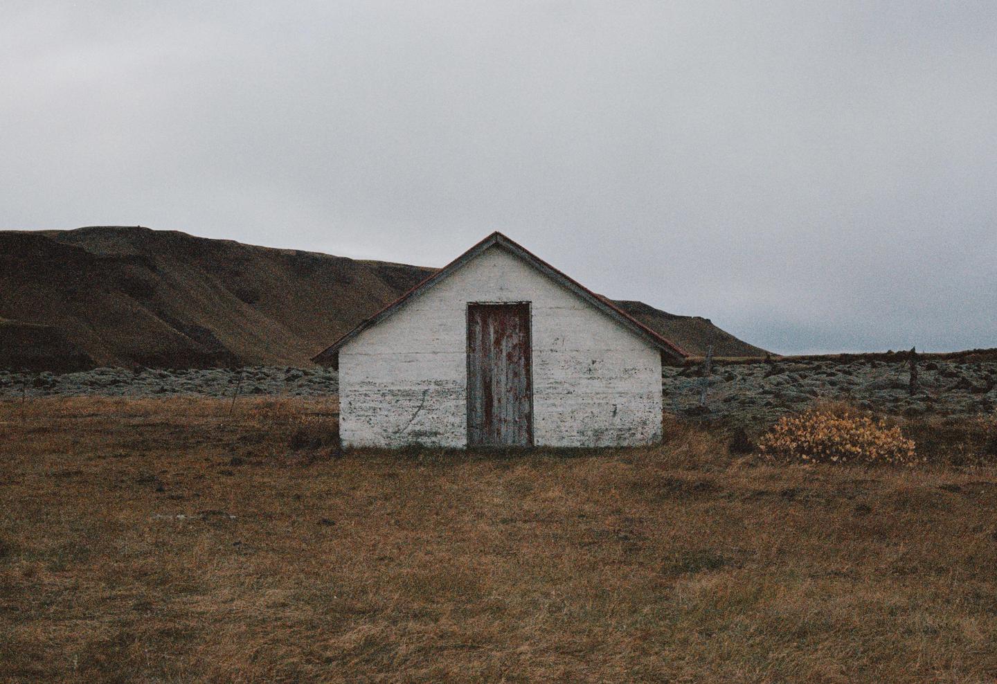 A small white shed with a red door stands in a field with a mountain in the background under a cloudy sky.