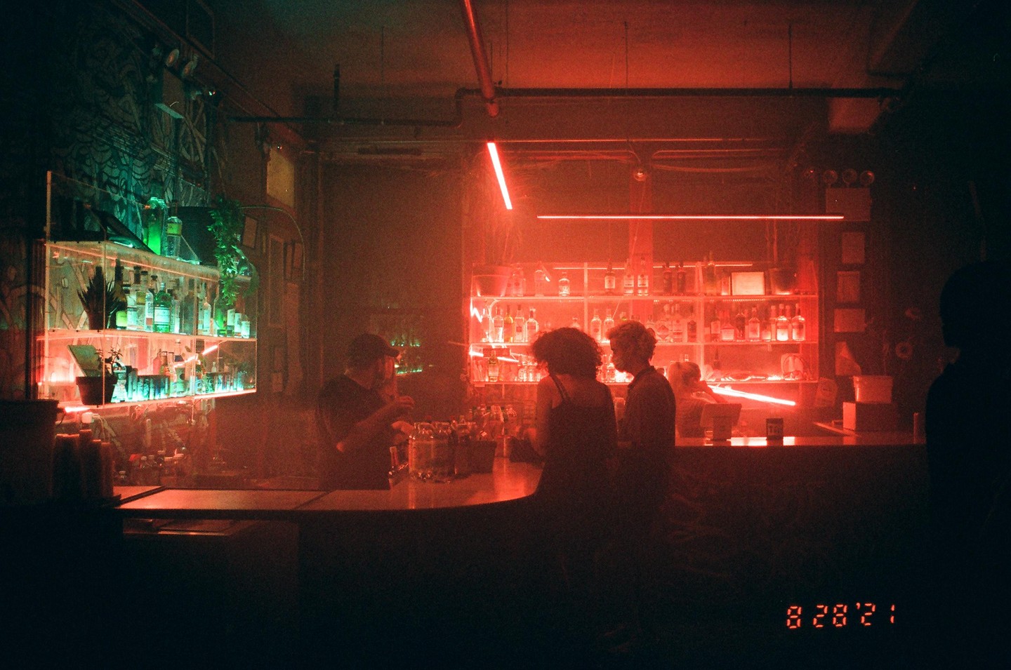 A bartender serves drinks to two people at a bar lit with red and green neon lights.